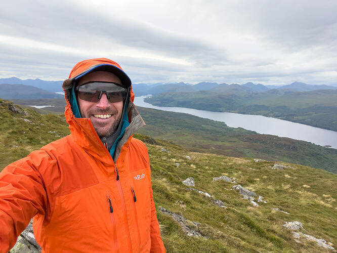 Wearing lots of layers at the summit, with Loch Katrine behind